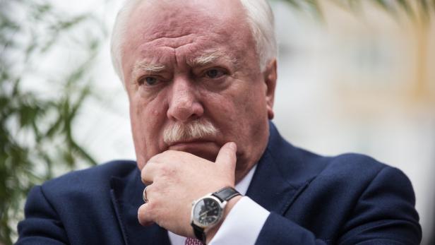 Ein Mann mit weißem Haar und Schnurrbart in einem dunklen Anzug denkt nach.