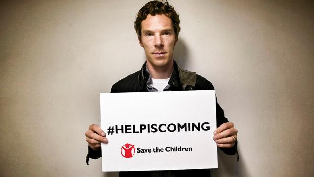 Benedict Cumberbatch hält ein Schild mit der Aufschrift „#HELPISCOMING“ und dem Logo von Save the Children.
