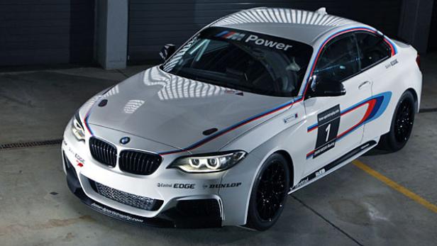 Ein weißer BMW M235i Rennwagen mit blauen und roten Streifen steht in einer Garage.