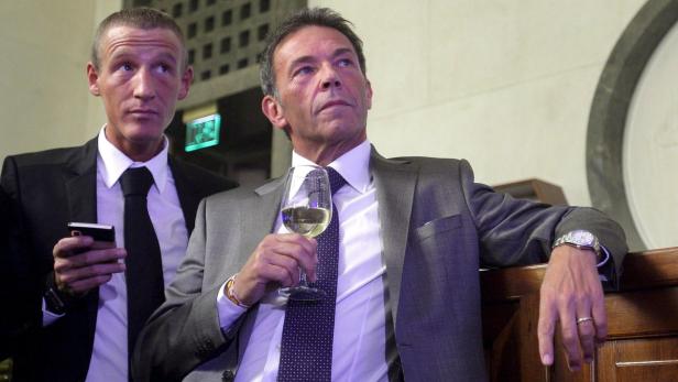 Zwei Männer in Anzügen, einer mit einem Glas Weißwein, der andere mit einem Smartphone.