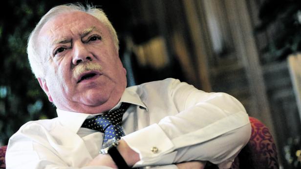 Ein älterer Mann mit Schnurrbart und Hemd mit Krawatte sitzt mit verschränkten Armen da.