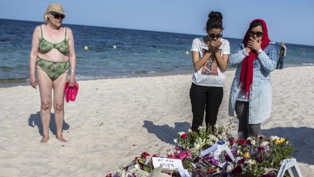 Zwei Frauen trauern an einem Blumenmonument am Strand, während eine ältere Frau im Badeanzug daneben steht.