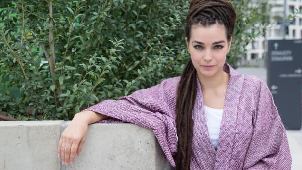 Eine Frau mit Dreadlocks und einem lila Kimono lehnt an einer Betonmauer.