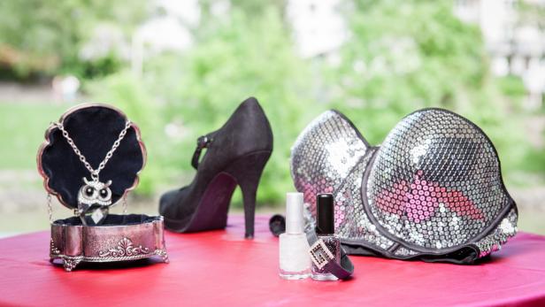 Ein Schmuckkästchen mit einer Eulen-Halskette, ein High Heel, Nagellack und ein Pailletten-BH auf einem Tisch.