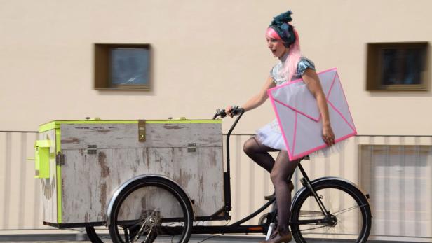 Eine Frau mit rosa Haaren fährt ein Lastenfahrrad und hält einen übergroßen Briefumschlag.