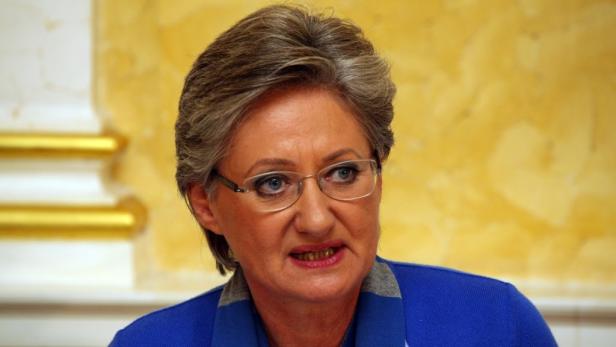 Eine Frau mit Brille und blauem Oberteil vor einem gelben Hintergrund.