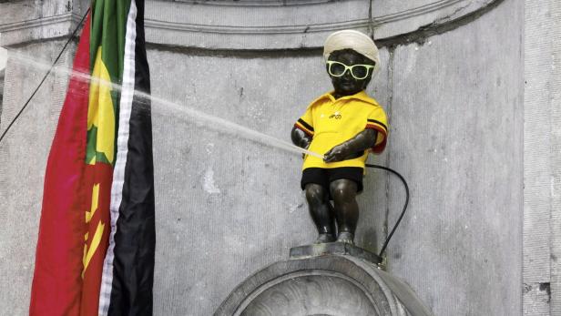 Die Manneken-Pis-Statue in Brüssel, verkleidet mit Hut, Sonnenbrille und Trikot, uriniert neben einer Flagge.