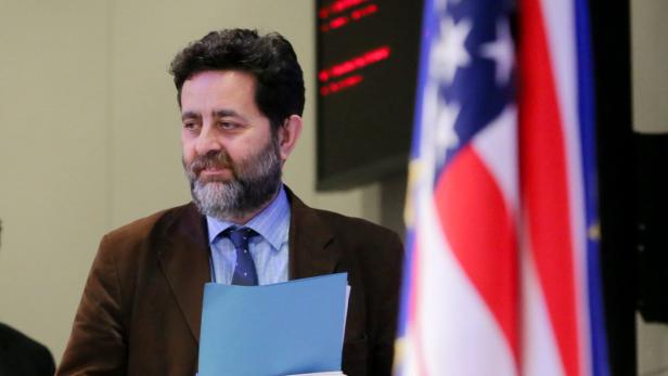 Ein Mann mit Bart hält eine blaue Mappe vor einer amerikanischen Flagge.