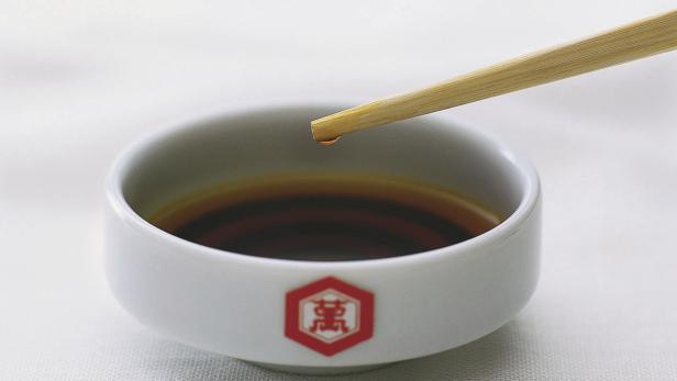 Ein Tropfen Sojasauce fällt von einem Essstäbchen in eine Schale.