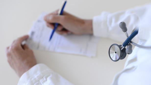 Ein Arzt schreibt mit einem Stift auf ein Dokument, ein Stethoskop liegt daneben.