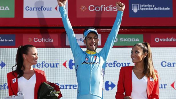 Ein Radrennfahrer in blauem Trikot jubelt auf dem Podium, flankiert von zwei Frauen.