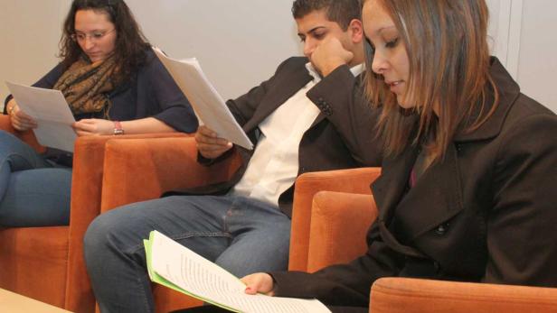 Drei Personen sitzen in orangefarbenen Sesseln und lesen Dokumente.