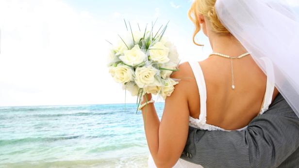 Eine Braut mit einem Rosenstrauß wird von einem Mann am Strand umarmt.