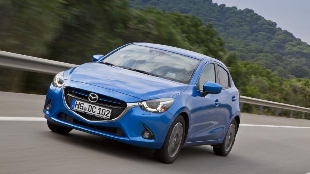 Ein blauer Mazda fährt auf einer Landstraße an einem bewaldeten Hügel vorbei.