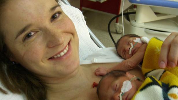Eine lächelnde Frau hält zwei Neugeborene im Arm, die mit Sauerstoffschläuchen versorgt werden.