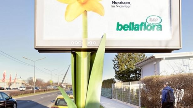 Eine Werbetafel in Form einer Narzisse wirbt für „bellaflora“ mit dem Slogan „Frühling!“.