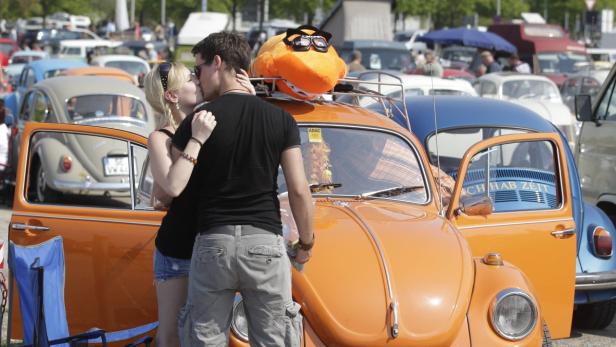 Ein Paar küsst sich vor einem orangefarbenen VW Käfer mit einem Plüschtier auf dem Dach.