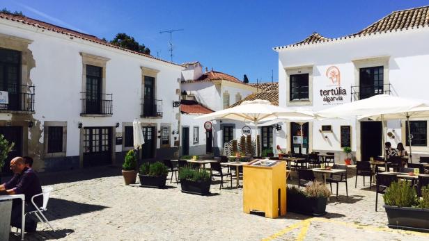 Ein Platz mit Tischen und Stühlen vor weißen Gebäuden mit dem Schriftzug „Tertúlia Algarvia“.