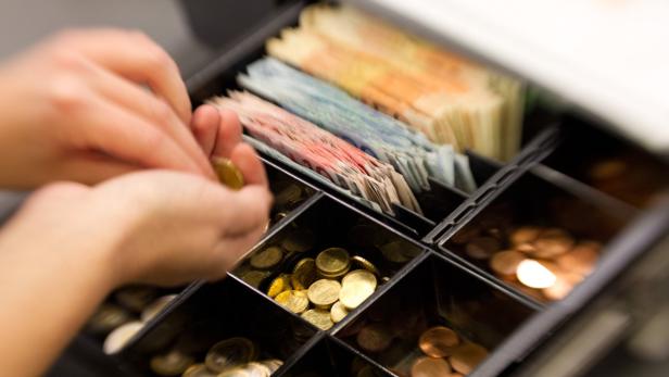 Eine Person hantiert mit Münzen vor einer offenen Kasse mit Geldscheinen und Münzgeld.