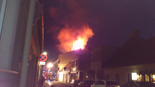 In einer Straße steht ein Gebäude in Flammen, während Feuerwehrleute eintreffen.