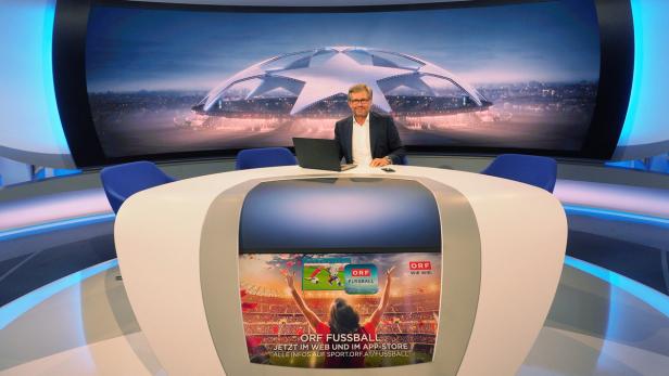 Ein Mann sitzt in einem Fernsehstudio vor einem Bildschirm mit einem Fußballstadion.