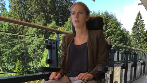 Eine junge Frau sitzt in einem Rollstuhl auf einem Balkon mit Blick auf Bäume.