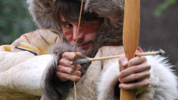 Ein Mann mit Gesichtsbemalung zielt mit Pfeil und Bogen.