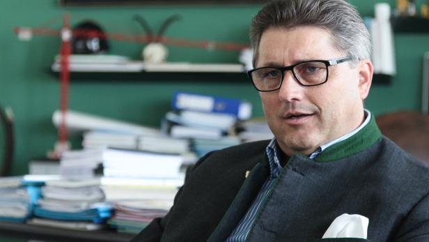 Ein Mann mit Brille und Trachtenjacke sitzt vor einem Schreibtisch mit Bauplänen und einem Kranmodell.