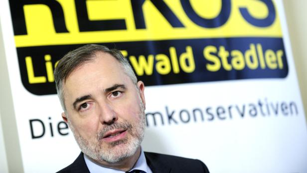 Ein Mann mit Bart steht vor einem Schild mit der Aufschrift „Die Liberalen und Konservativen“.