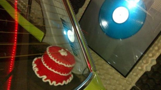 Ein gehäkelter roter Kuchen und eine blaue Schallplatte sind in einem Auto zu sehen.