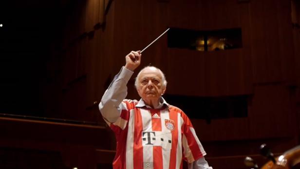 Ein Dirigent im Bayern-München-Trikot dirigiert ein Orchester.