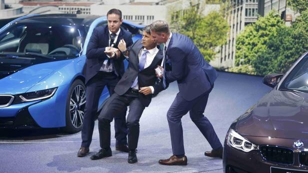 Zwei Männer in Anzügen führen eine dritte Person ab, im Hintergrund sind BMW-Fahrzeuge zu sehen.