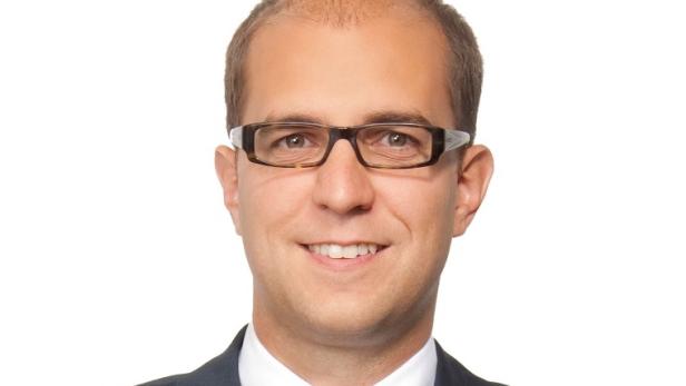 Ein lächelnder Mann mit Brille und Anzug vor einem weißen Hintergrund.