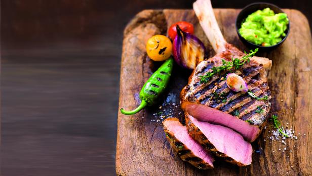 Ein Tomahawk-Steak mit gegrilltem Gemüse und Guacamole auf einem Holzbrett.