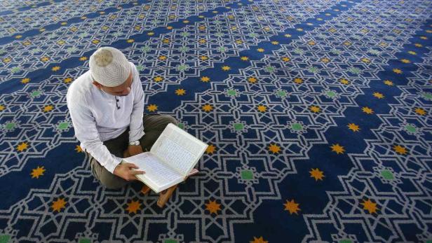 Ein Mann mit Kappe liest den Koran auf einem verzierten Teppich.