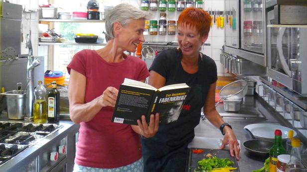 Zwei Frauen stehen in einer Küche und betrachten ein Buch von Eva Rossmann mit dem Titel „Fadenkreuz“.