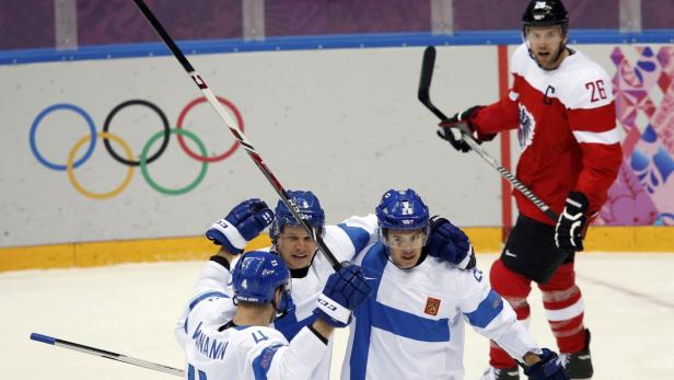 Finnische Hockeyspieler feiern vor dem Hintergrund der olympischen Ringe ein Tor gegen Österreich.