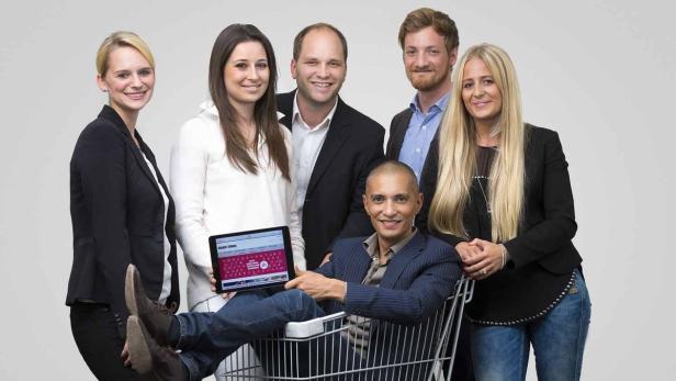 Eine Gruppe von sechs Personen posiert, wobei ein Mann in einem Einkaufswagen sitzt und ein Tablet hält.