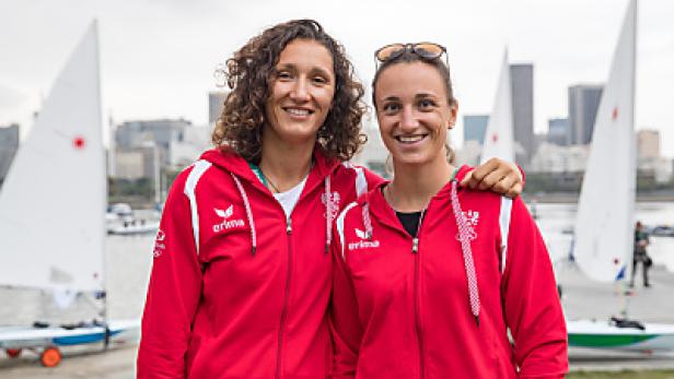 Zwei Seglerinnen in roter Trainingskleidung posieren vor Segelbooten und einer Stadtkulisse.