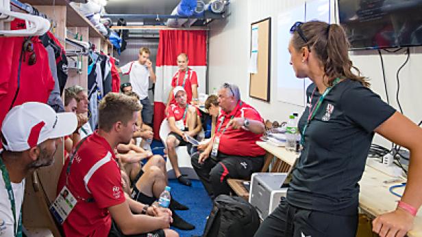 Eine Gruppe von Sportlern und Trainern bespricht eine Strategie in einem kleinen Raum.