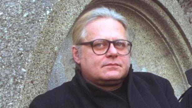 Ein Mann mit Brille und dunklem Mantel vor einer steinernen Wand.