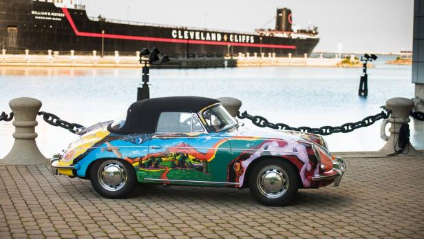 Ein bunt bemalter Porsche steht vor einem See mit einem Schiff der Cleveland Cliffs Reederei im Hintergrund.