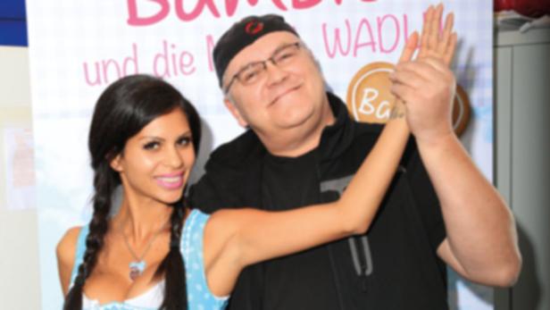 Eine Frau im Dirndl und ein Mann mit Brille posieren vor einer Werbetafel.