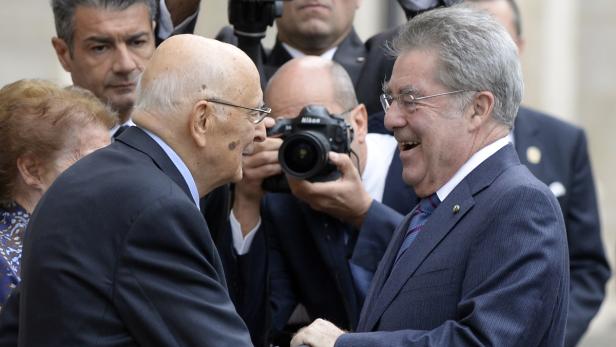 Zwei ältere Männer in Anzügen schütteln sich die Hände, während sie von Fotografen umgeben sind.