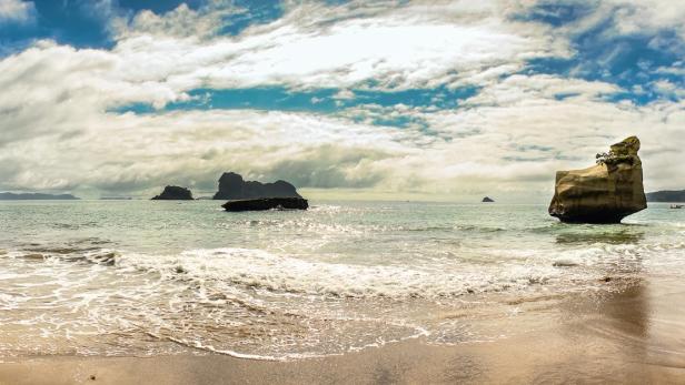 Panoramaaufnahme eines Strandes mit Felseninseln im Meer unter einem bewölkten Himmel.