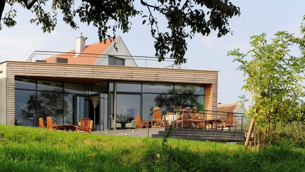 Modernes Haus mit großen Glasfronten, Holzverkleidung und Terrasse mit Holzmöbeln, umgeben von grünem Rasen und Bäumen.