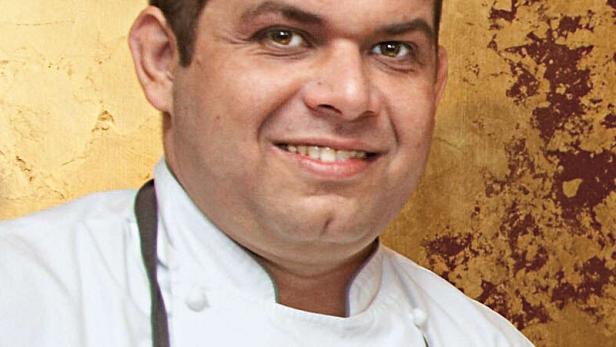 Ein lächelnder Koch in Kochuniform vor einem goldenen Hintergrund.