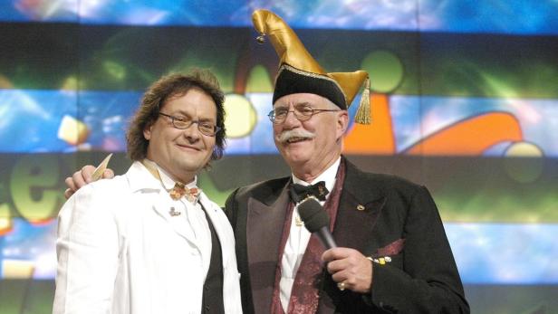 Zwei Männer, einer mit Narrenkappe und Mikrofon, stehen auf einer Bühne.