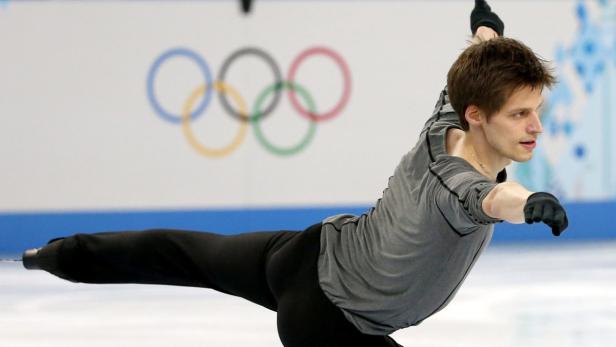 Ein Eiskunstläufer zeigt eine Pirouette vor dem Hintergrund der olympischen Ringe.
