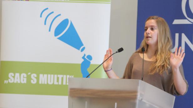 Eine Frau hält eine Präsentation hinter einem Rednerpult mit dem Slogan „Sag’s Multi!“.